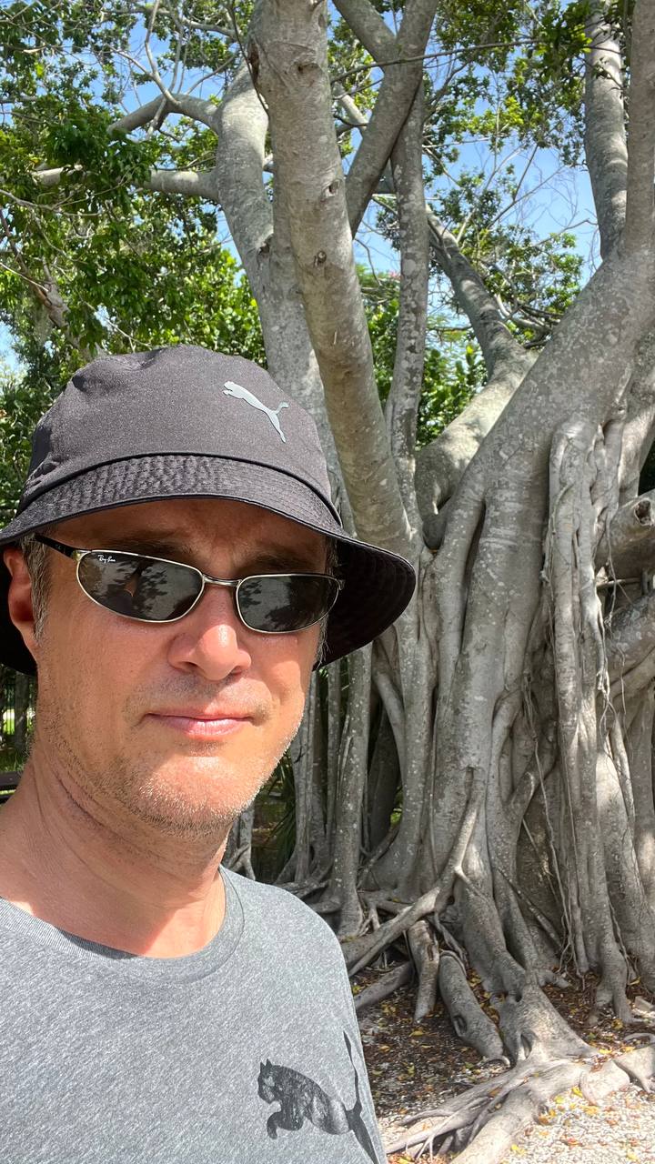 Mann mit Puma-Bucket-Hut vor einem großen Baum mit dicken Wurzeln im Robinson Preserve Park